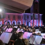 WhatsApp Image 2026-08-04 at 41236 PM 4 - عروبة الاخباري | Oroba News Large outdoor orchestra at night with a conductor directing musicians, colorful building lights in red, blue, and purple in the background.