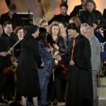 WhatsApp Image 2026-08-04 at 41237 PM 1 - عروبة الاخباري | Oroba News Bishop in ceremonial robes shakes hands with a woman in a blue evening dress on stage, surrounded by an orchestra with musicians and violins nearby