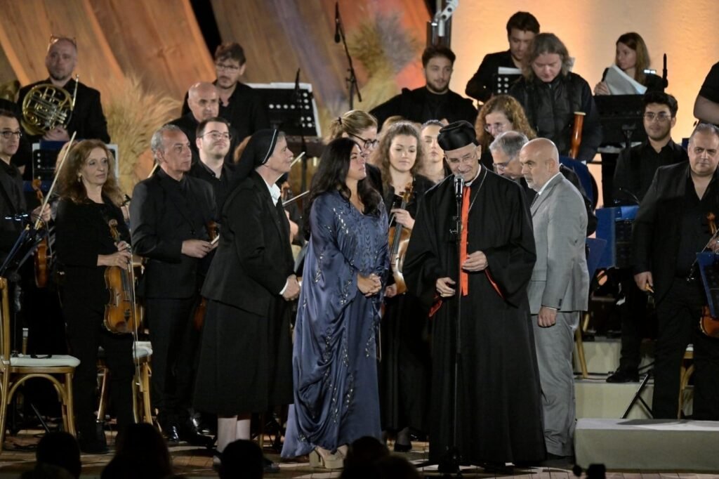 WhatsApp Image 2026-08-04 at 41237 PM - عروبة الاخباري | Oroba News Religious leader in black robes with an orange stole speaks into a microphone on stage, surrounded by a seated orchestra and formally dressed attendees.