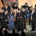 WhatsApp Image 2026-08-04 at 41237 PM 2 - عروبة الاخباري | Oroba News Conductor in black robe with red trim speaks into a microphone on stage, surrounded by an orchestra in formal attire.