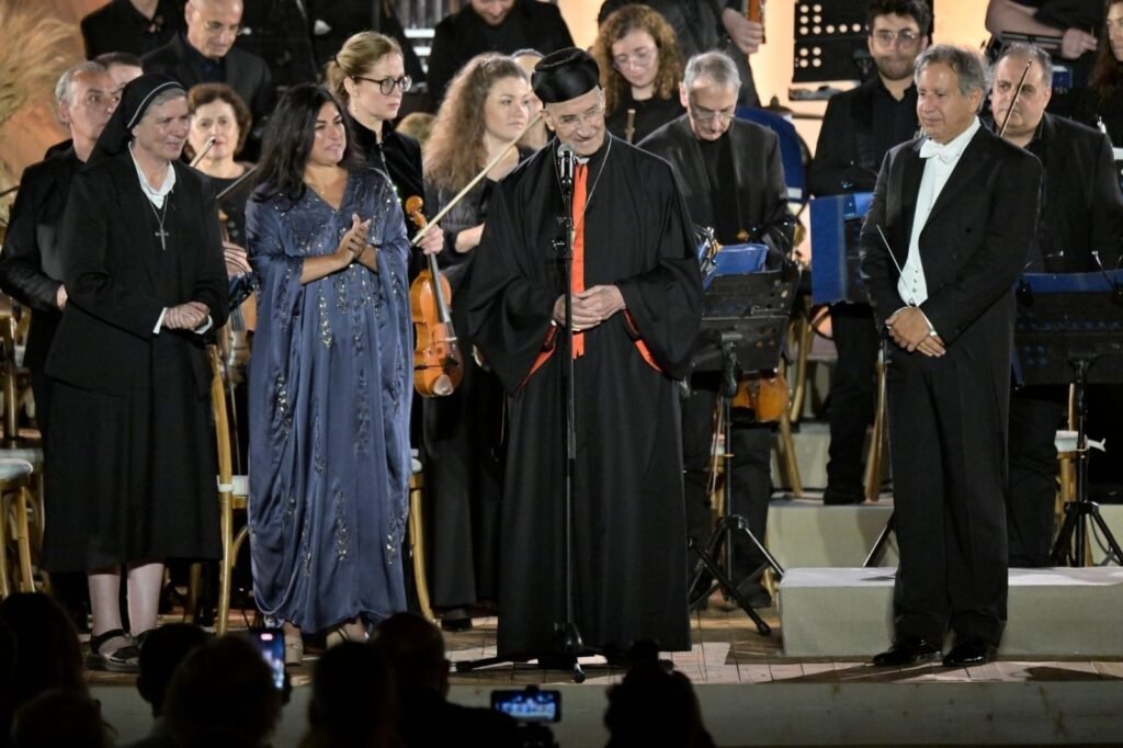 WhatsApp Image 2026-08-04 at 41237 PM 3 - عروبة الاخباري | Oroba News Man in ceremonial black robe with red tie speaks at a microphone on stage, surrounded by musicians and a nun in a head covering behind him.