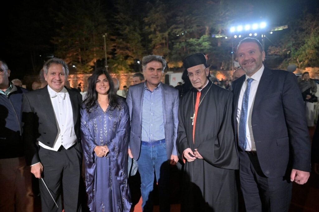 WhatsApp Image 2026-08-04 at 41237 PM 6 - عروبة الاخباري | Oroba News Group of five adults posing for a photo at an outdoor evening event, with two men in suits, a woman in a blue gown, a man in a blazer, and a clergyman in black robes with a red tie, trees and bright event lights in the background.