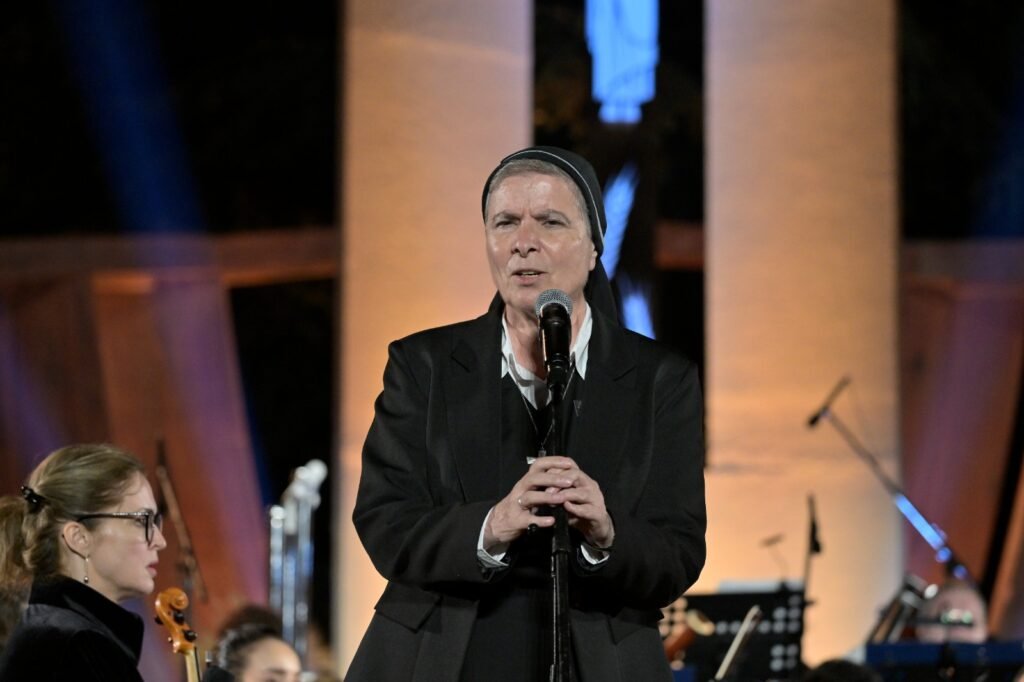 WhatsApp Image 2026-08-04 at 41238 PM - عروبة الاخباري | Oroba News Woman in a dark suit and head covering sings into a microphone on a stage with an orchestra in the background.