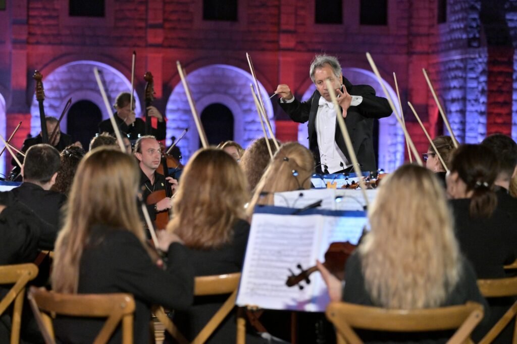 WhatsApp Image 2026-08-04 at 41238 PM 2 - عروبة الاخباري | Oroba News Conductor in a tuxedo leads an orchestra on stage, baton raised while musicians play string instruments amid red‑blue architectural lighting.