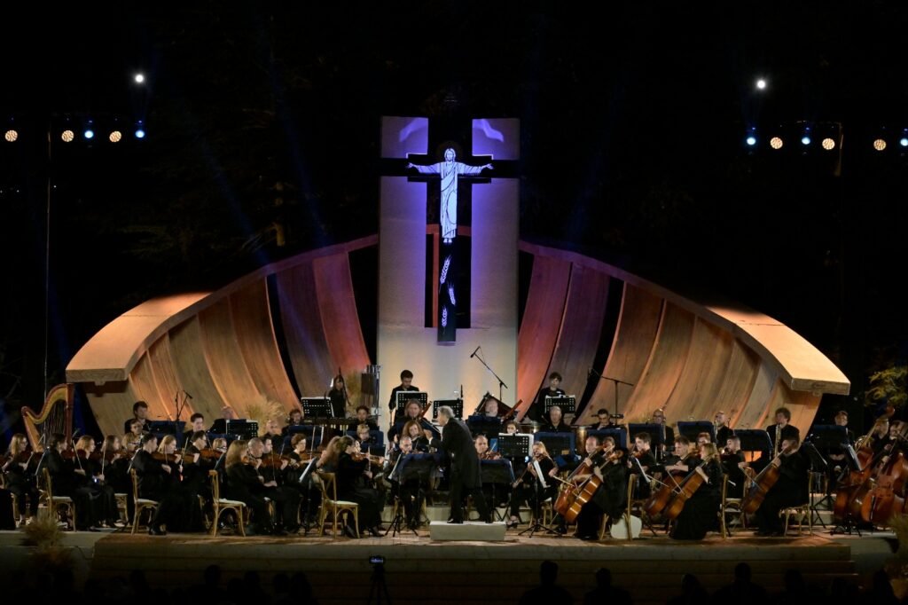 WhatsApp Image 2026-08-04 at 41238 PM 3 - عروبة الاخباري | Oroba News Outdoor night concert: full symphony orchestra with conductor at center, lit stage against curved wooden backdrop and cross sculpture behind.