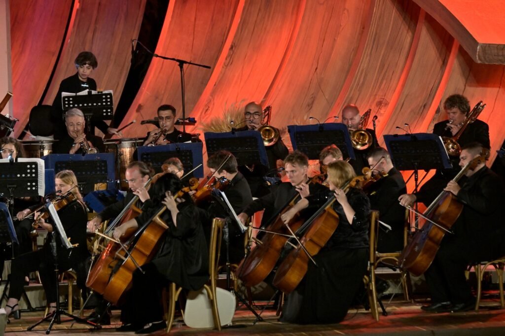 WhatsApp Image 2026-08-04 at 41238 PM 4 - عروبة الاخباري | Oroba News Full symphony orchestra performing on stage in black attire, with string players in front and brass behind, against an orange curved-wall backdrop.