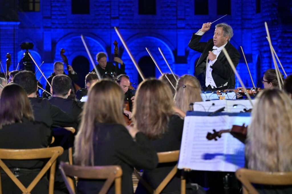 WhatsApp Image 2026-08-04 at 41238 PM 5 - عروبة الاخباري | Oroba News Conductor in a tuxedo raises baton while leading a symphony orchestra on a blue-lit stage, with string players in the foreground and sheet music visible.