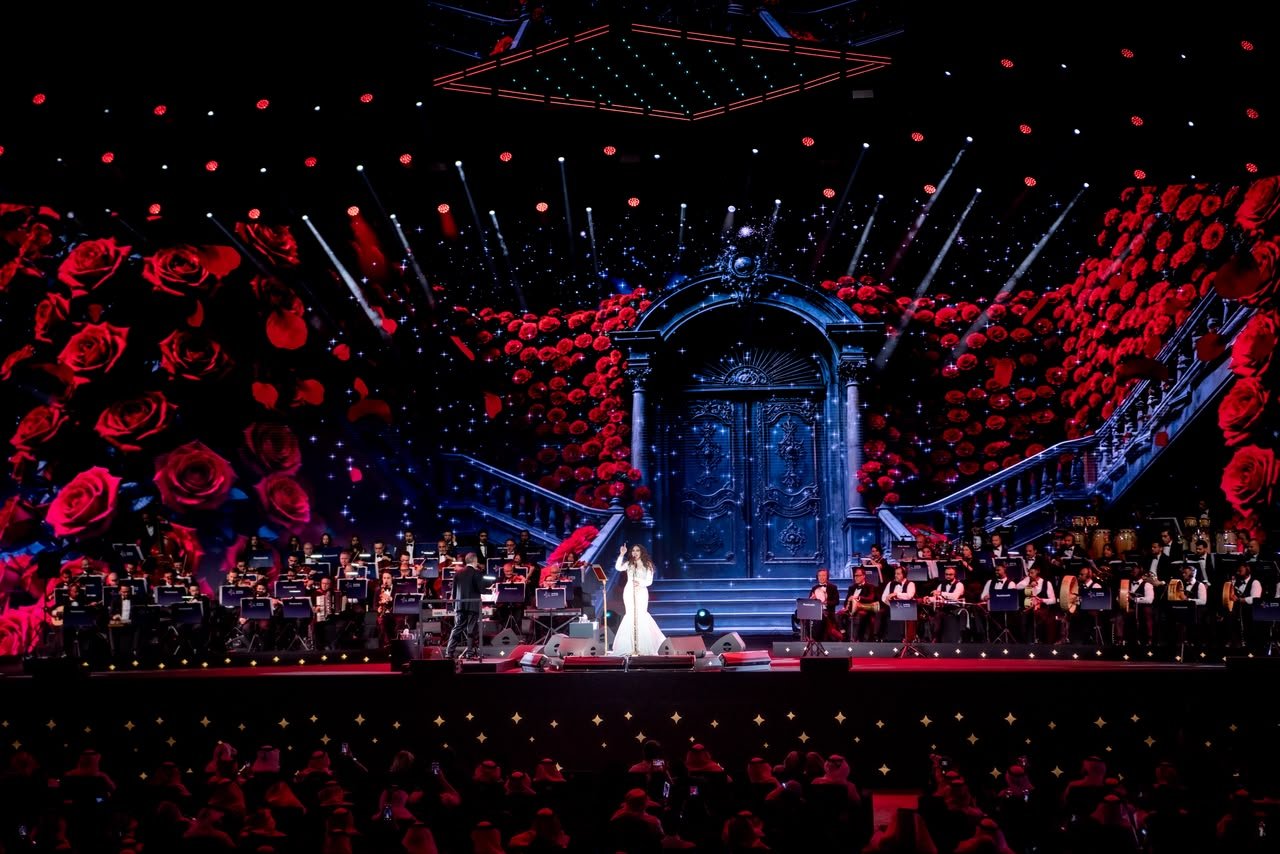 WhatsApp Image 2026-08-04 at 80705 PM Singer in a white gown centers stage with a full orchestra, against a backdrop of red roses and an ornate door scene at a grand concert.