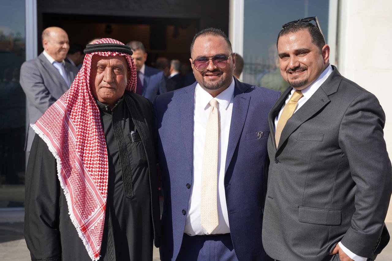 WhatsApp Image 2026-08-05 at 32452 PM - عروبة الاخباري | Oroba News Three men in formal attire posing outdoors, center man in a blue suit and pale yellow tie flanked by a man in traditional red‑and‑white keffiyeh and a man in a gray suit.