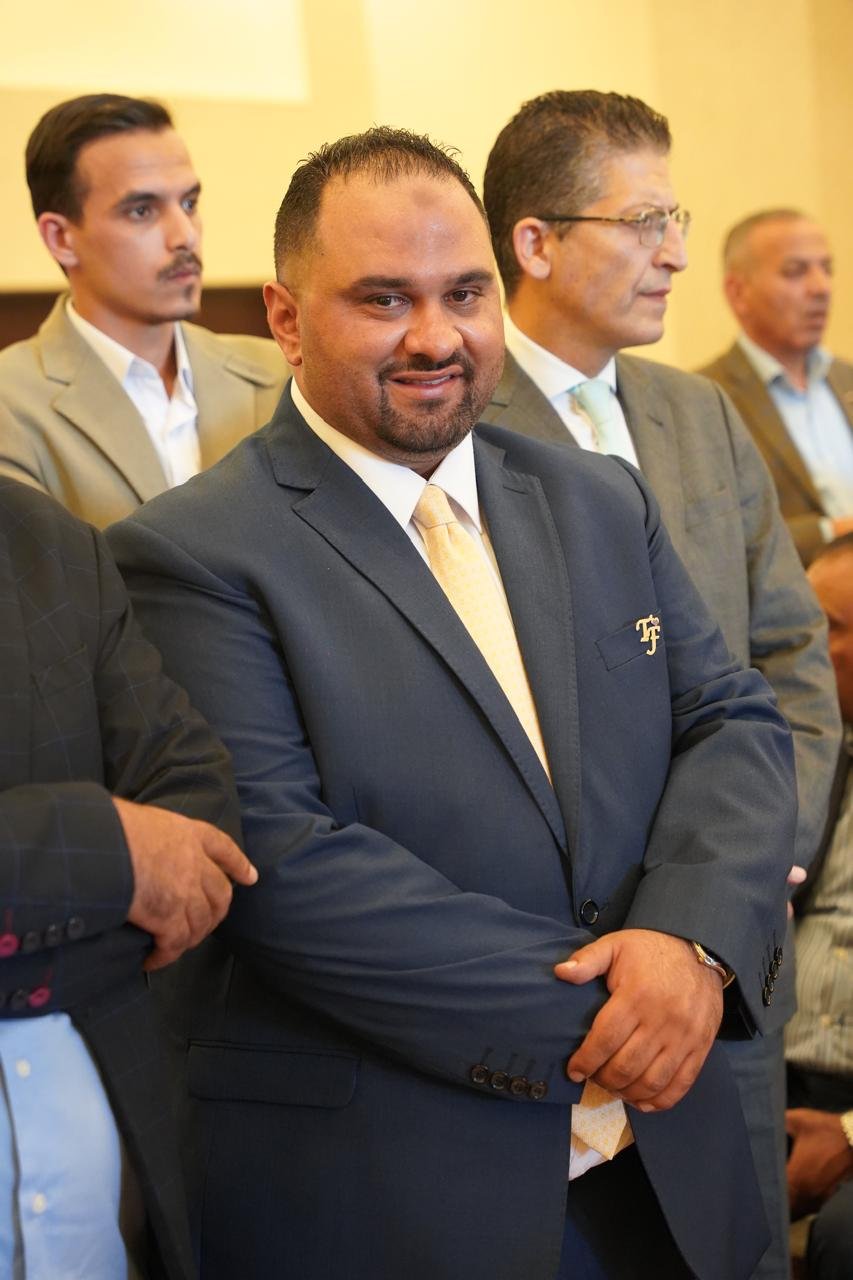 WhatsApp Image 2026-08-05 at 32452 PM2 - عروبة الاخباري | Oroba News Smiling man in a navy suit and pale yellow tie at a formal event, with other suited men in the background.