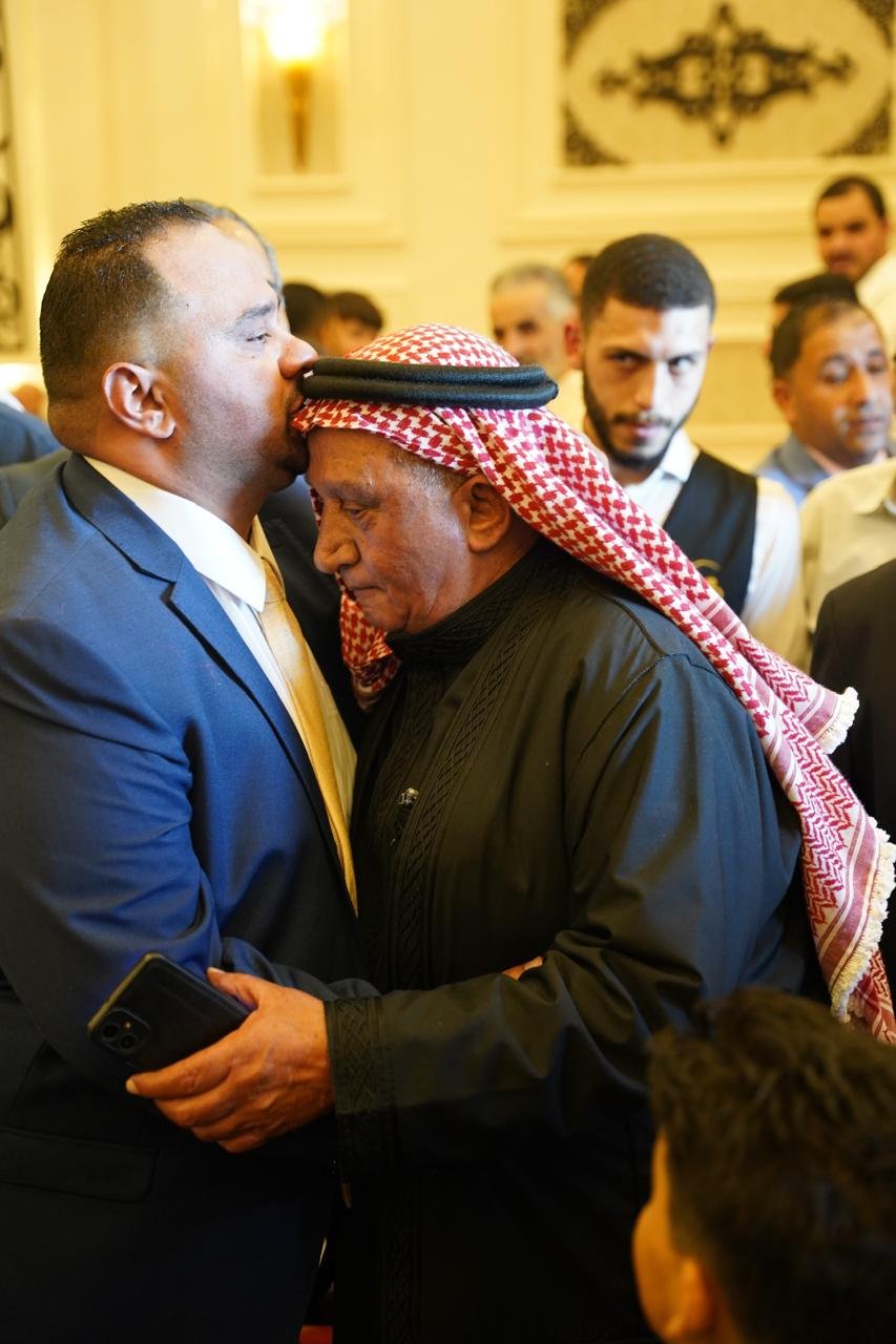 WhatsApp Image 2026-08-05 at 32452 PM3 - عروبة الاخباري | Oroba News Man in a suit gently kisses an older man’s forehead at a formal gathering, both wearing traditional attire, with others in the background.