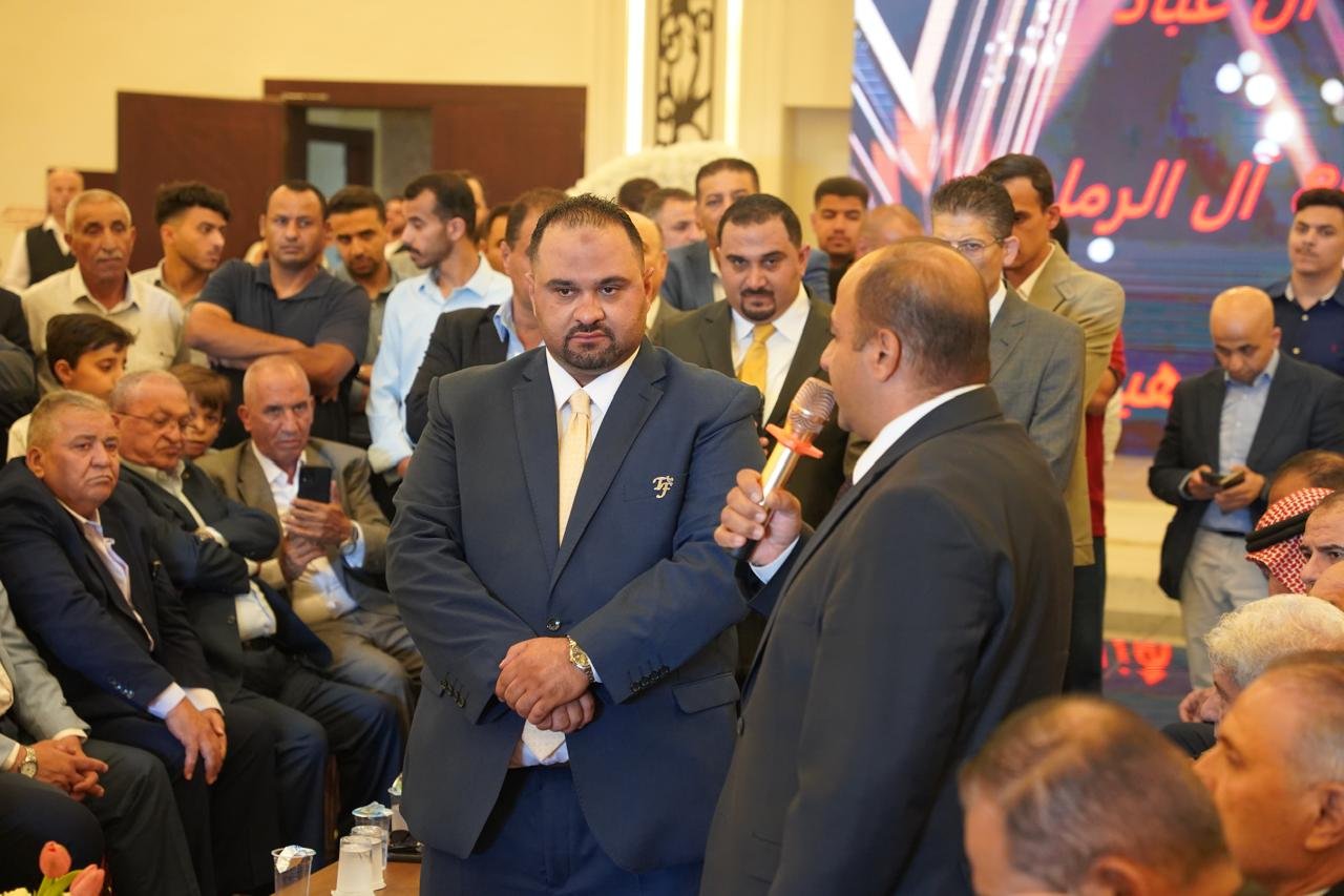 WhatsApp Image 2026-08-05 at 32452 PM4 - عروبة الاخباري | Oroba News Two men in suits on a stage at a formal gathering; one speaks into a microphone while the other stands with arms crossed in front of a crowded audience.