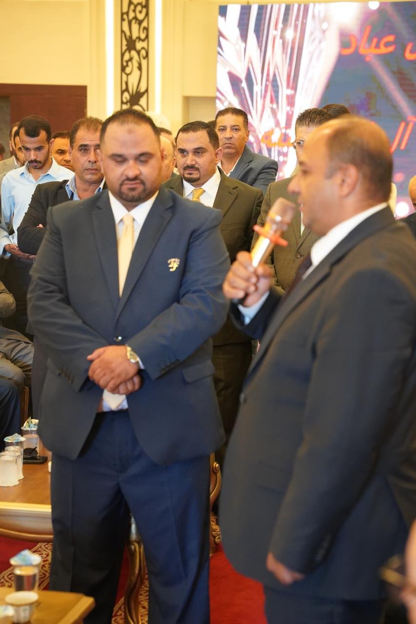 WhatsApp Image 2026-08-05 at 32453 PM - عروبة الاخباري | Oroba News Man in a dark suit holds a microphone while another man in a blue suit stands nearby at a formal event with several suited attendees in the background.