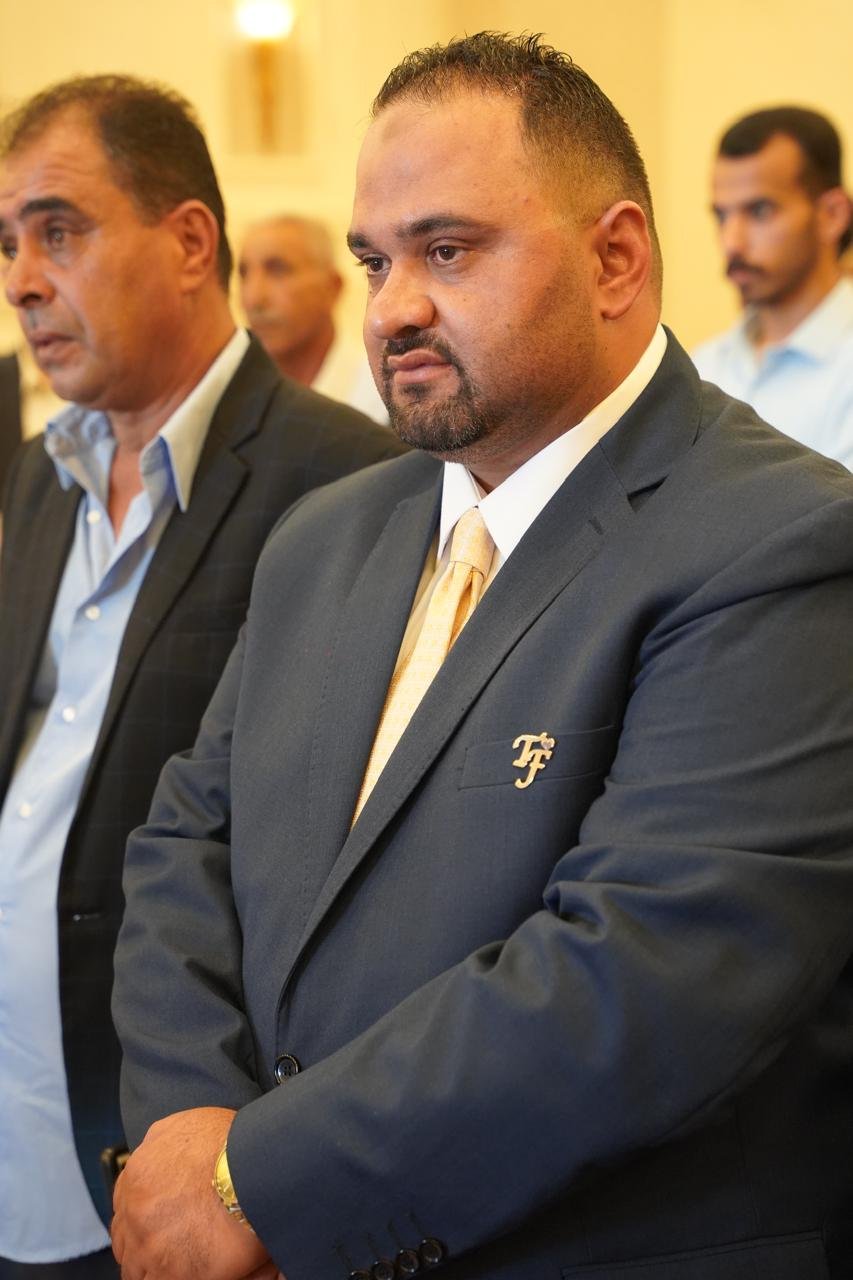 WhatsApp Image 2026-08-05 at 32453 PM2 - عروبة الاخباري | Oroba News Man in a dark suit with a gold tie stands with folded arms at a formal event; other attendees are blurred in the background.