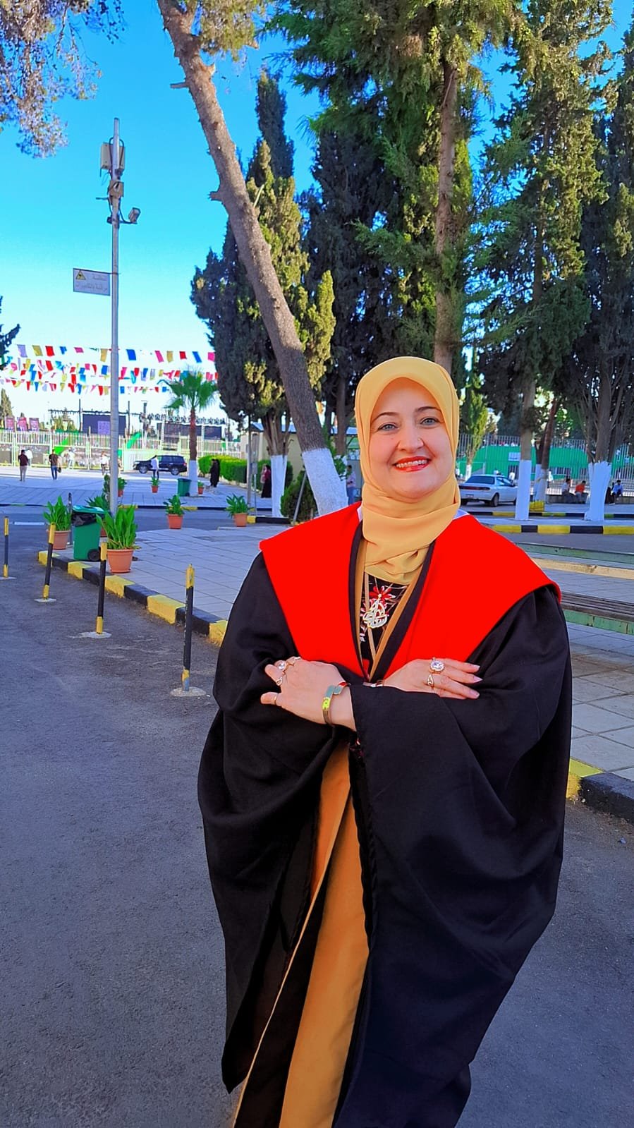 Smiling woman in a black graduation gown with a red stole and yellow hijab, standing outdoors with trees in the background and arms crossed.