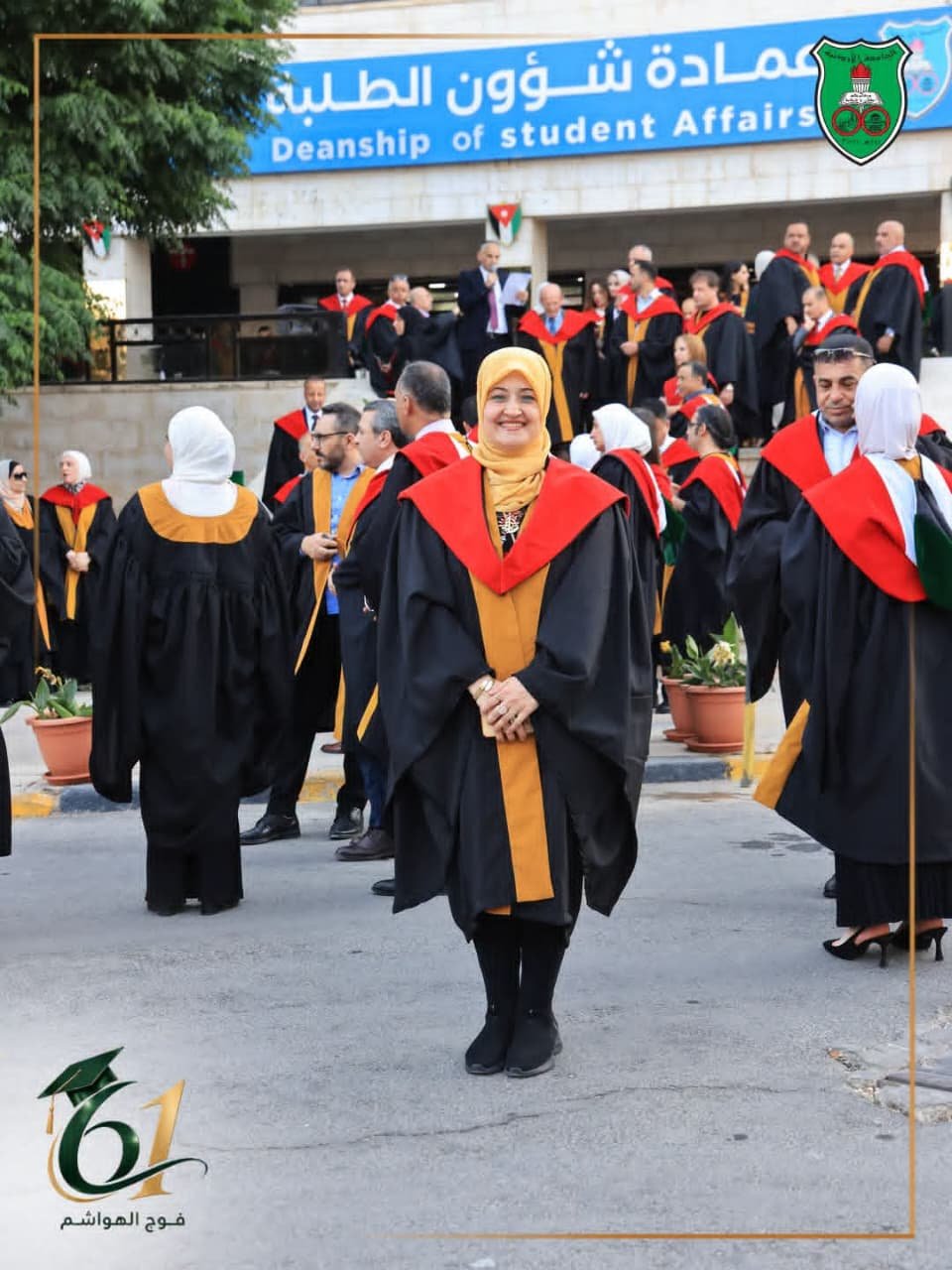 Smiling graduate in a black gown with red and gold trim and a yellow hijab stands among other graduates at an outdoor university ceremony outside a building with a blue Deanship of Student Affairs sign.