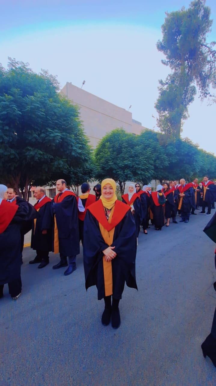 Smiling graduate in a yellow hijab leading a line of graduates in black gowns with red-and-gold sashes outdoors on a street.