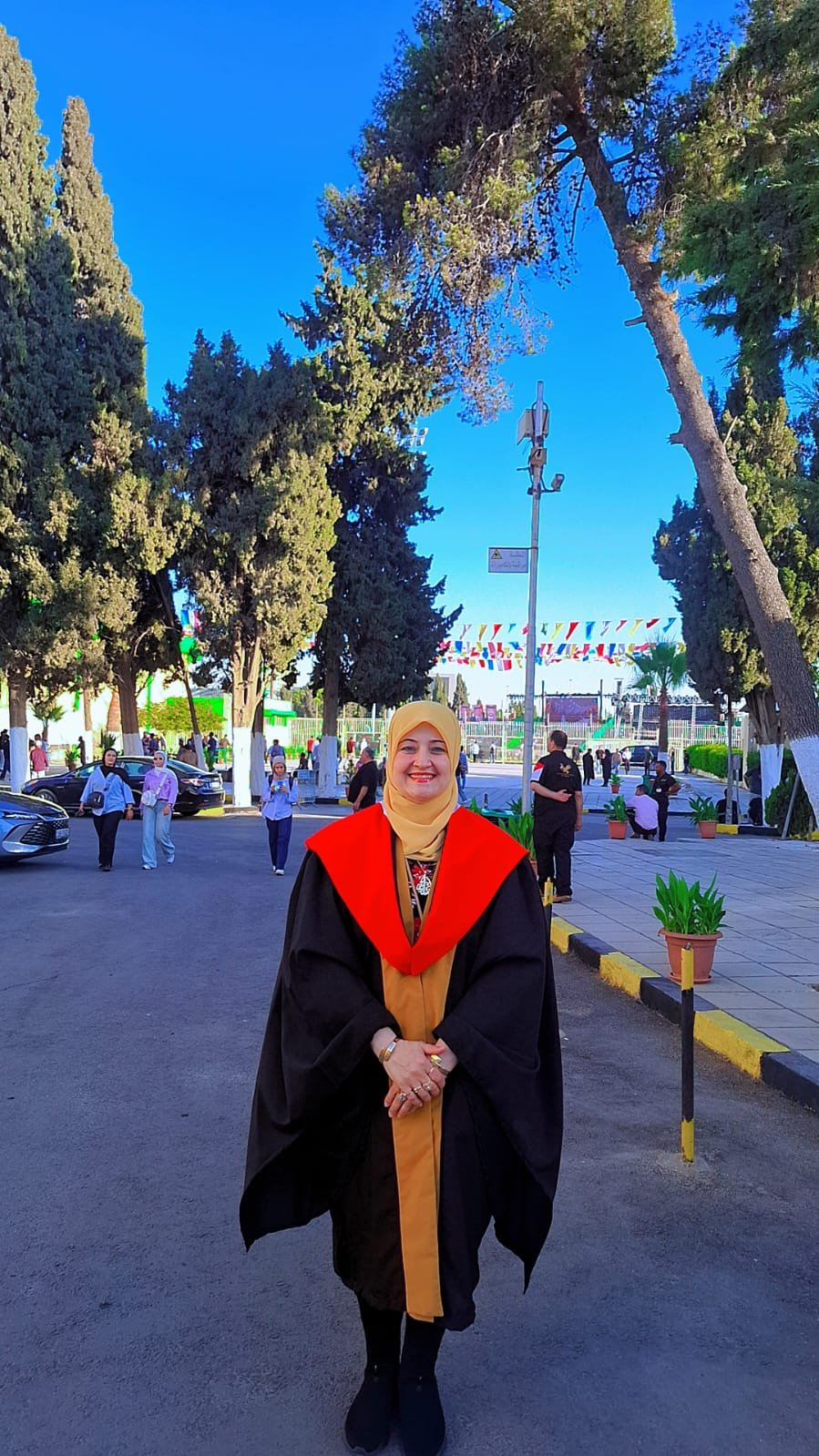 Graduate in a black gown with a red and gold hood stands smiling on a sunny street at an outdoor ceremony.