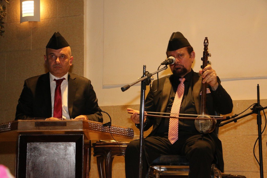 WhatsApp Image 2026-08-06 at 41349 PM - عروبة الاخباري | Oroba News Two men in suits and hats perform on stage: one plays a harmonium, the other plays a stringed instrument with a microphone nearby.