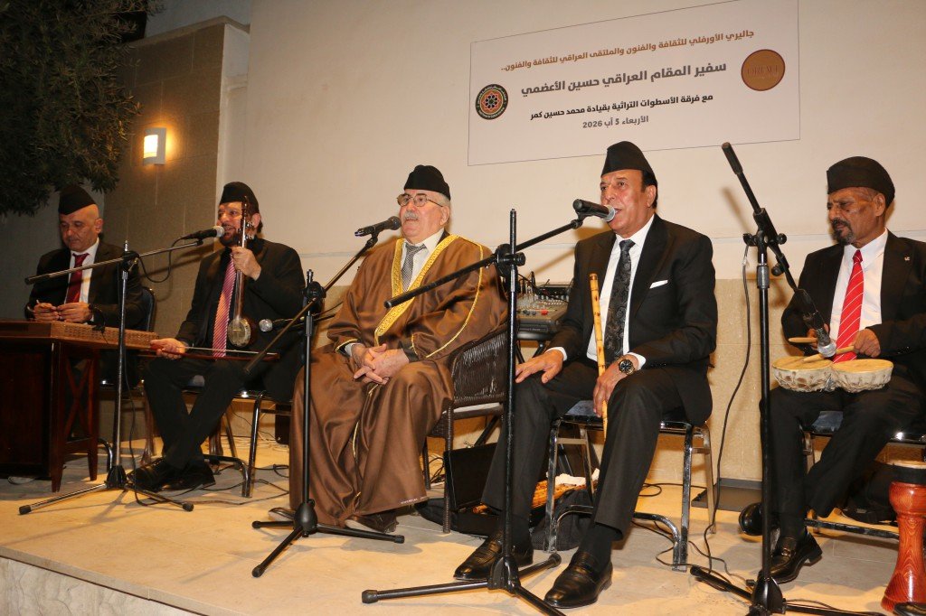 WhatsApp Image 2026-08-06 at 41350 PM - عروبة الاخباري | Oroba News Six men in traditional robes and suits sit on a stage with microphones, performing at a cultural event, with an Arabic banner on the wall behind them.