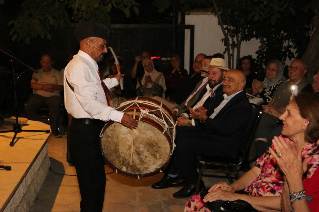 WhatsApp Image 2026-08-06 at 41353 PM - عروبة الاخباري | Oroba News Drummer in a white shirt and black hat plays a large frame drum to an attentive, seated outdoor audience.