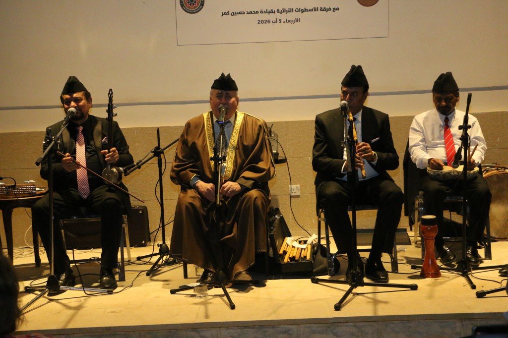 WhatsApp Image 2026-08-06 at 41354 PM - عروبة الاخباري | Oroba News Four men in traditional and formal attire sit on a stage with microphones, performing music; central figure wears a brown robe with gold trim while others wear suits and fezzes.