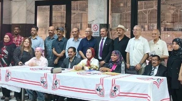 Group of people posing for a formal photo around a long table with white cloth and red logos, some seated and others standing in a hall.