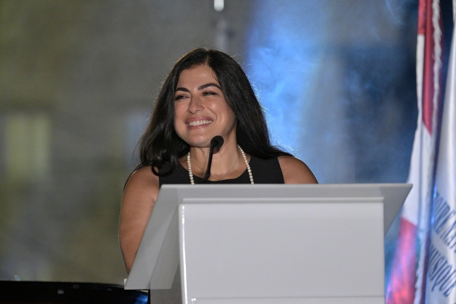 WhatsApp Image 2026-08-07 at 14939 PM - عروبة الاخباري | Oroba News Smiling woman in a black dress with a pearl necklace speaking at a white podium with a microphone, blue smoky background behind him/her.