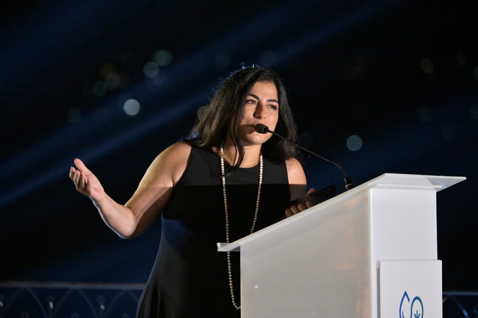 WhatsApp Image 2026-08-07 at 14941 PM - عروبة الاخباري | Oroba News Woman in a black dress with a pearl necklace speaks at a white podium on a dark stage with microphone present.