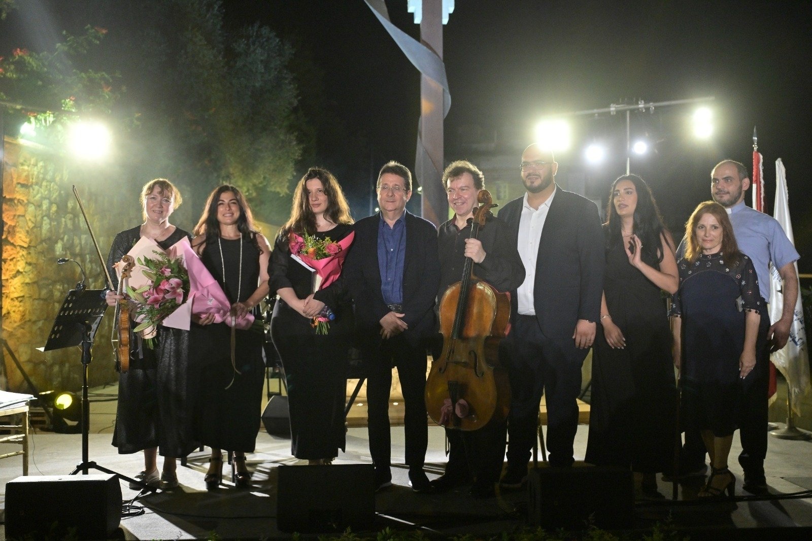 WhatsApp Image 2026-08-07 at 14943 PM 2 - عروبة الاخباري | Oroba News Group of performers on an outdoor stage at night, holding bouquets and instruments (violin, cello), posing for a photo.
