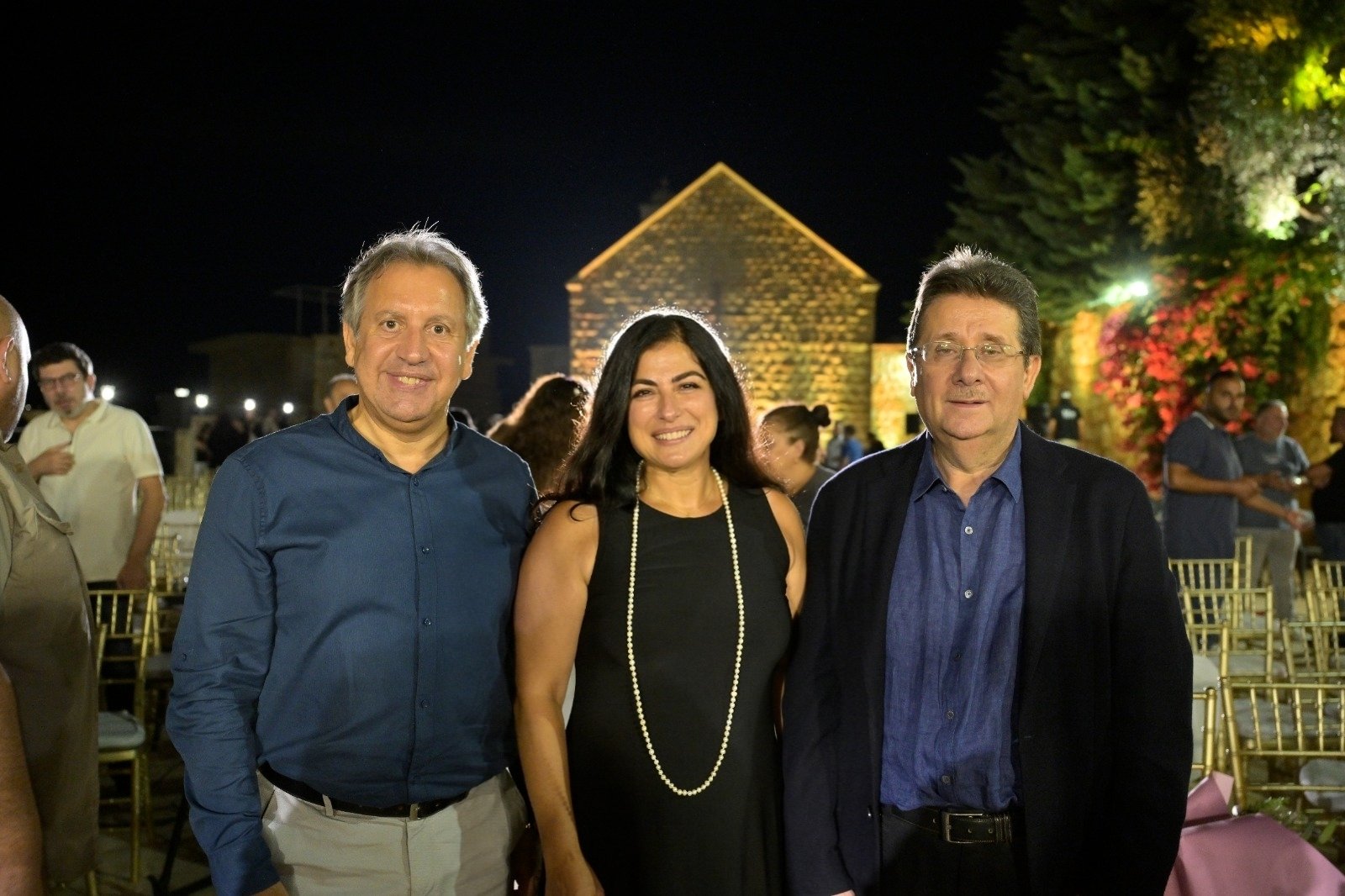 WhatsApp Image 2026-08-07 at 14943 PM 3 - عروبة الاخباري | Oroba News Three people pose and smile at a nighttime outdoor event, with a stone building and trees in the background.