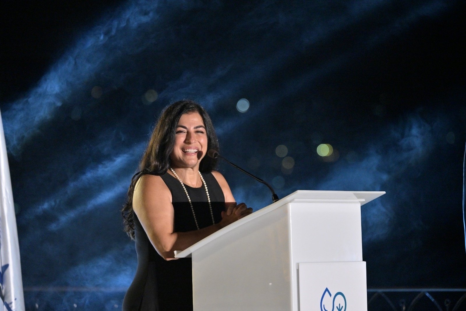 WhatsApp Image 2026-08-07 at 14944 PM 3 - عروبة الاخباري | Oroba News Woman in a black dress with a pearl necklace smiles while speaking at a white podium on a lit stage at night.