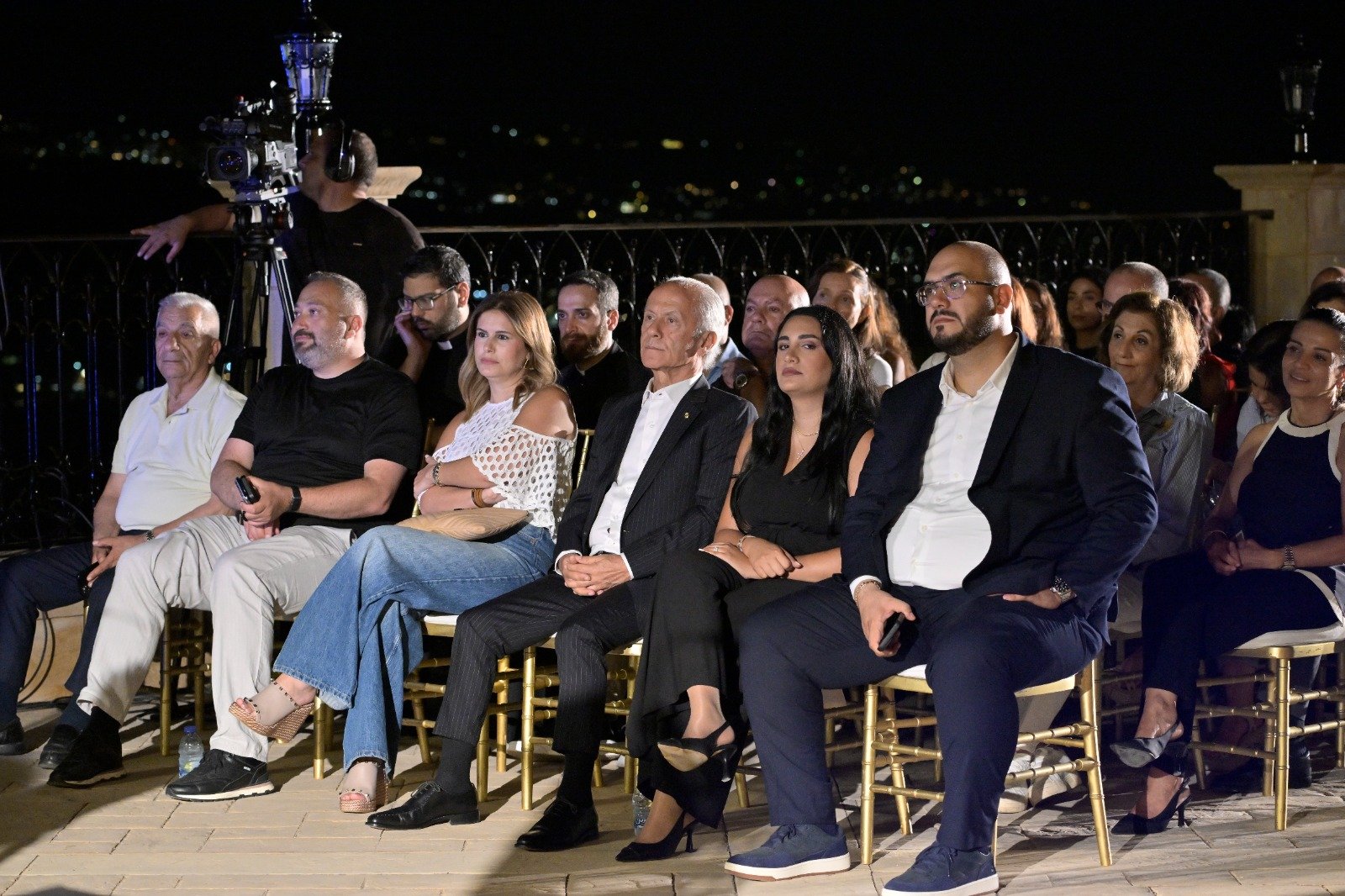 WhatsApp Image 2026-08-07 at 14945 PM 2 - عروبة الاخباري | Oroba News Audience seated outdoors at night watching a presentation; a camera on a tripod looms to the left.