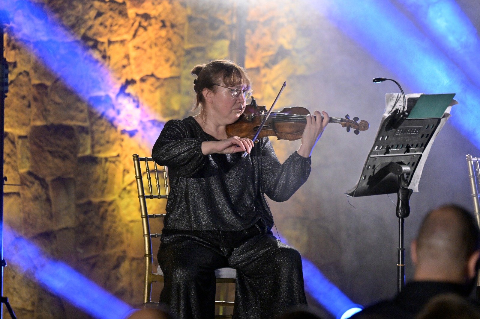 WhatsApp Image 2026-08-07 at 14946 PM 2 - عروبة الاخباري | Oroba News Female violinist in a glittery black outfit playing a violin on stage with a music stand nearby and blue and amber stage lighting.