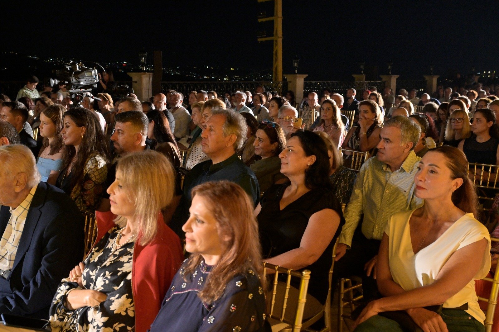 WhatsApp Image 2026-08-07 at 14946 PM 3 - عروبة الاخباري | Oroba News Crowd seated at an outdoor nighttime event, facing a stage with a camera on a tripod visible at the left.