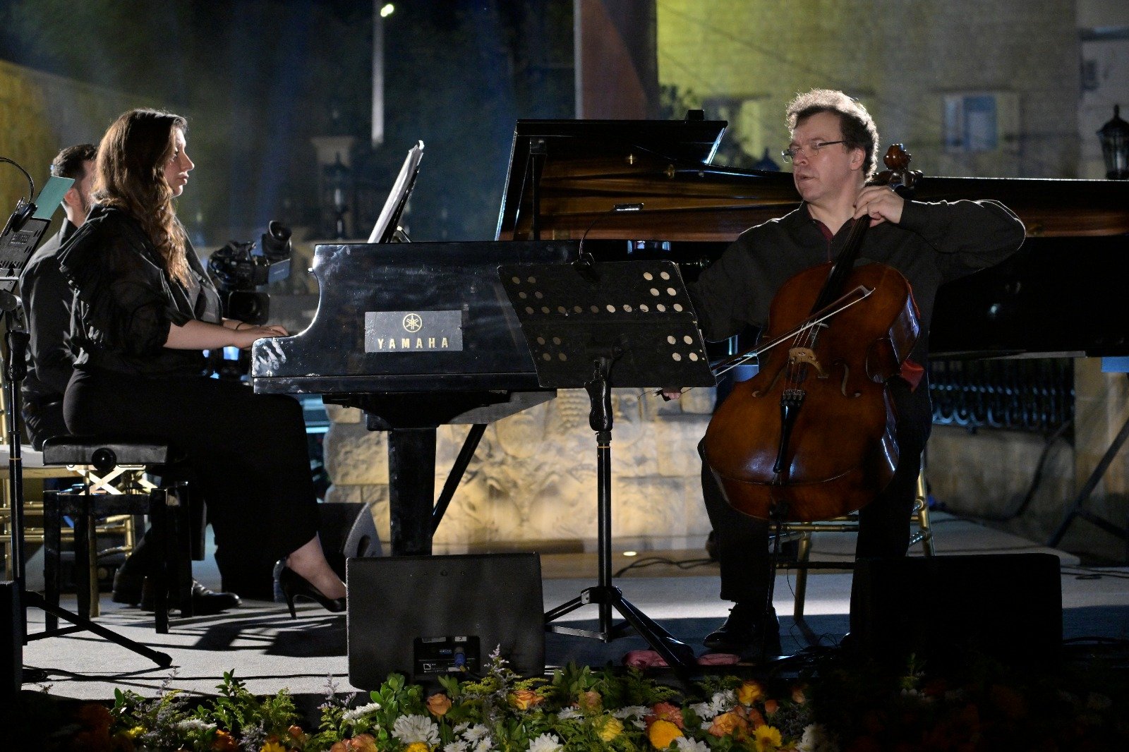 WhatsApp Image 2026-08-07 at 14946 PM 4 - عروبة الاخباري | Oroba News Pianist and cellist performing on an outdoor stage at night, with a Yamaha grand piano and sheet music nearby, flowers in the foreground.