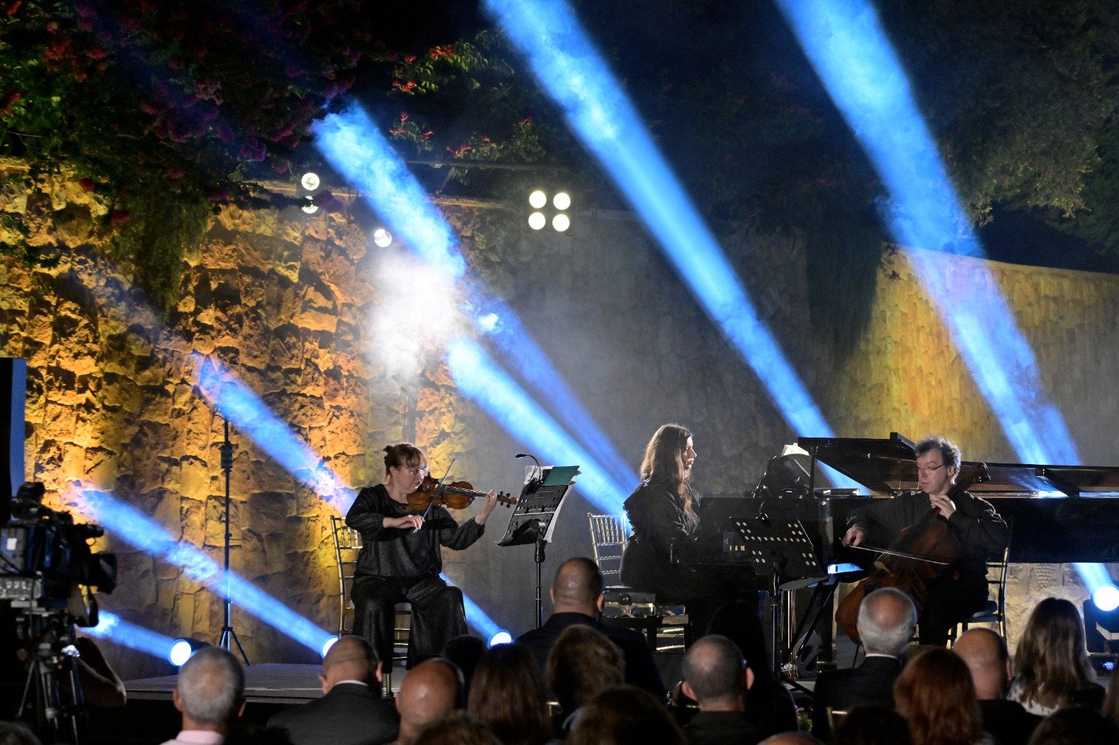 WhatsApp Image 2026-08-07 at 14946 PM - عروبة الاخباري | Oroba News Musicians perform on a stage: violinist, pianist, and cellist, with blue spotlights and a stone wall backdrop, audience watching at foreground.