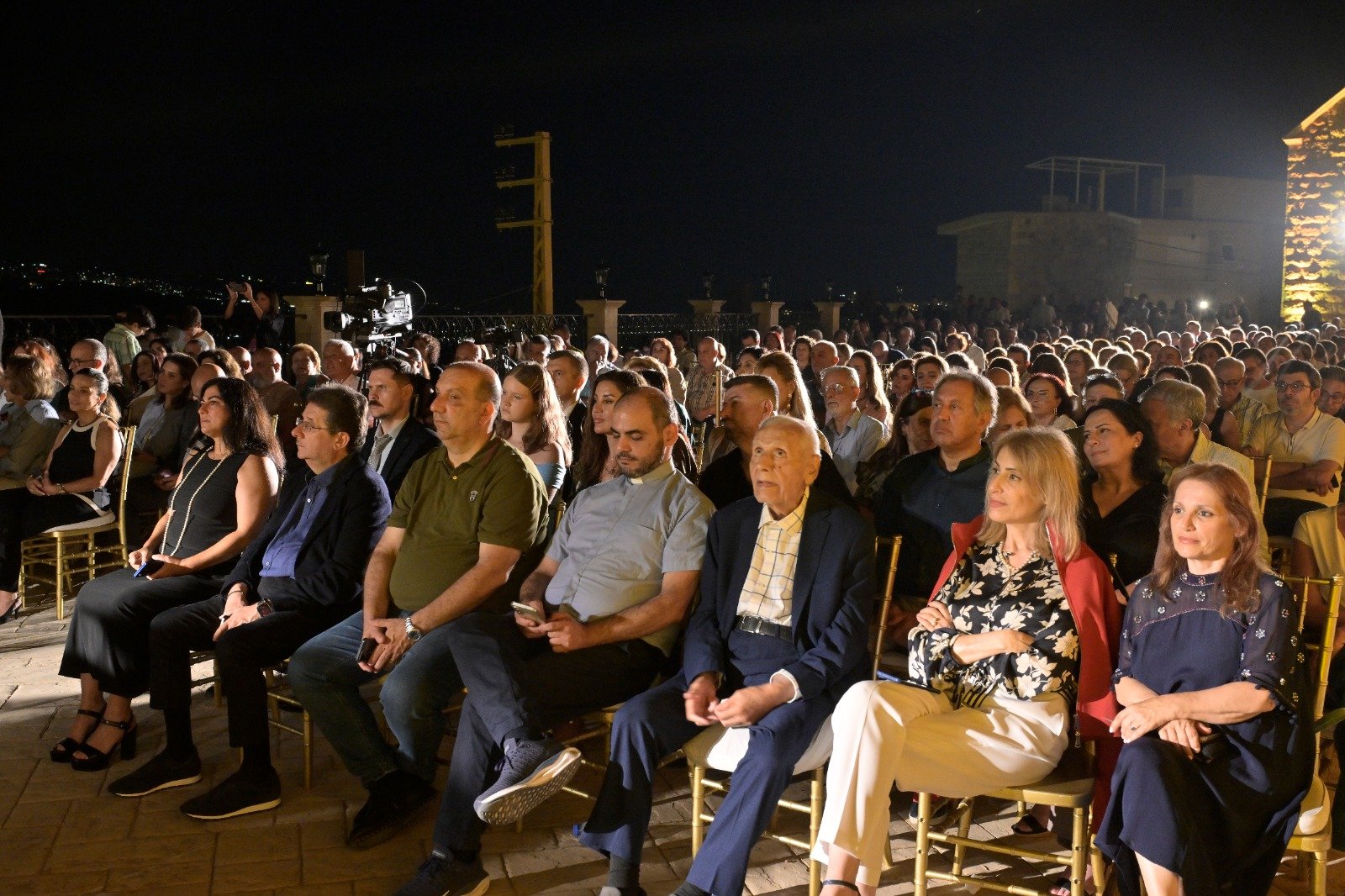 WhatsApp Image 2026-08-07 at 14947 PM 1 - عروبة الاخباري | Oroba News Large crowd seated at a nighttime outdoor event, with a camera and crew visible in the background nearby.