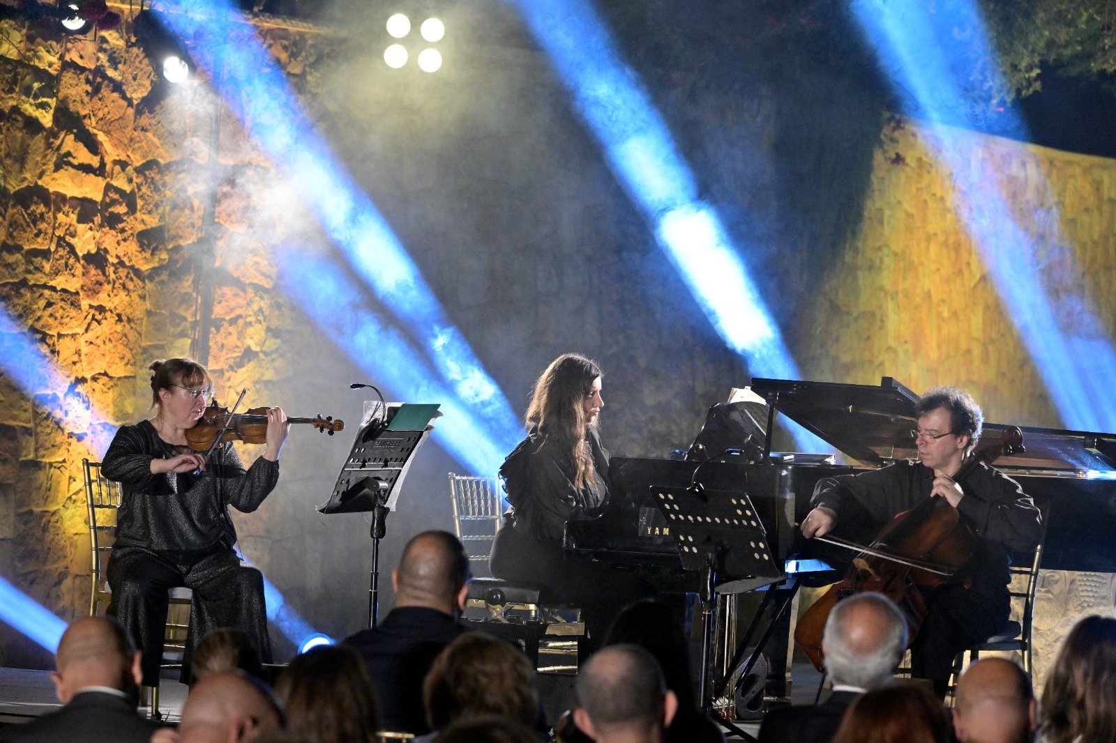 WhatsApp Image 2026-08-07 at 14947 PM 2 - عروبة الاخباري | Oroba News Violinist, pianist, and cellist performing on a stage with blue spotlight beams and a stone backdrop.