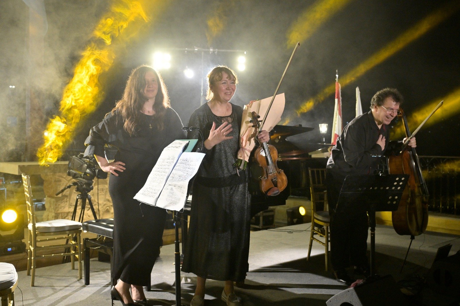 WhatsApp Image 2026-08-07 at 14947 PM - عروبة الاخباري | Oroba News Three musicians on a stage smiling after a performance: a violinist holding a bouquet beside two other players (one with sheet music) and a cellist, all in black with bright stage lighting in the background.