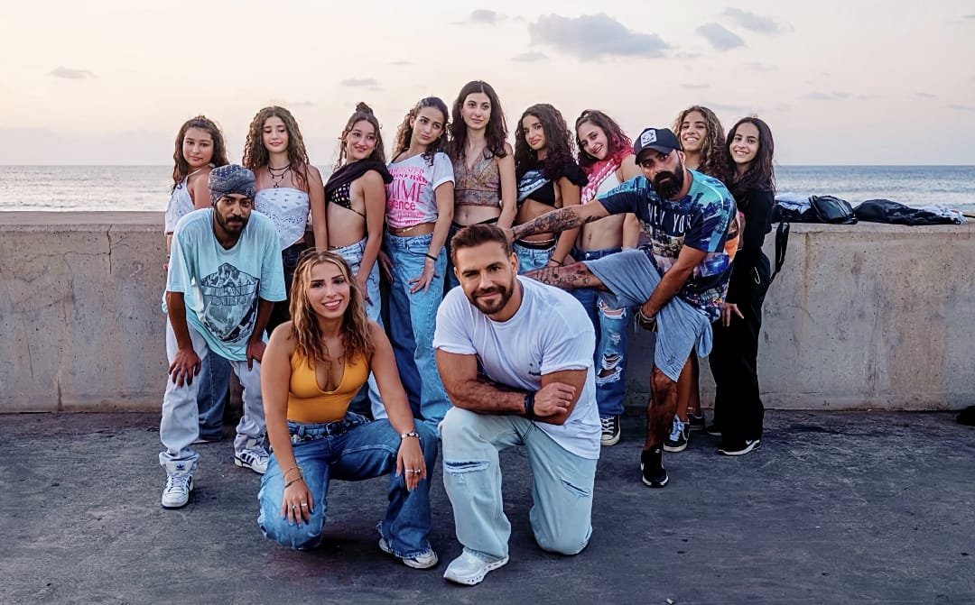 Group of diverse friends posing for a photo on a seaside boardwalk at sunset, with two individuals kneeling in front and others standing behind them.