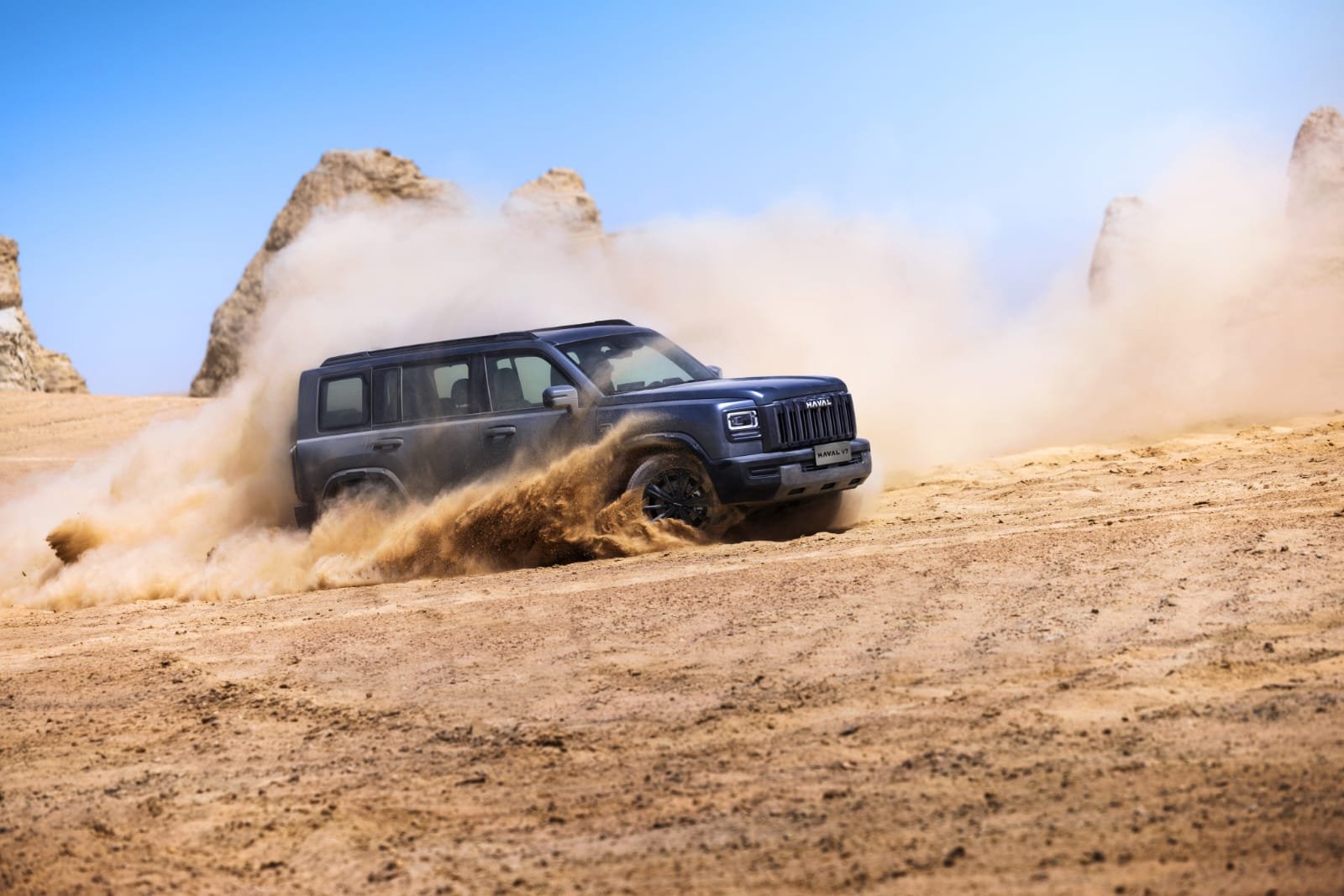 Gray off-road SUV kicking up a large dust cloud across a sandy desert, with rocky formations in the distance.