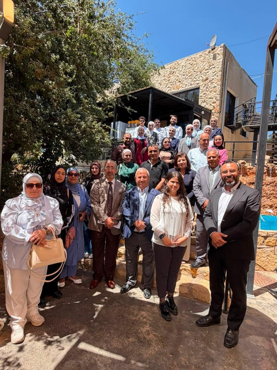 Group of men and women posing together outside a stone building on a sunny day, in a formal and casual mix of attire.