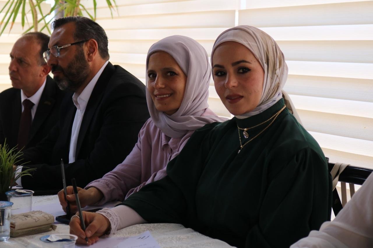 Two women in hijabs sit at a table signing documents, with two men beside them at a formal meeting table.