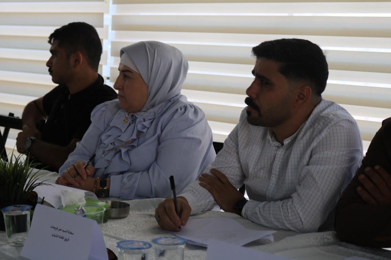 Three people sit at a conference table during a meeting; a woman in a light blue hijab writes on a notepad while a man in a white checkered shirt writes nearby, with another person on the left partially visible.