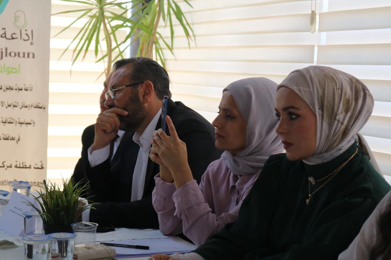 Three adults seated at a table in a bright room: a man in a suit with glasses and two women in hijabs, a banner with Arabic text visible on the left.