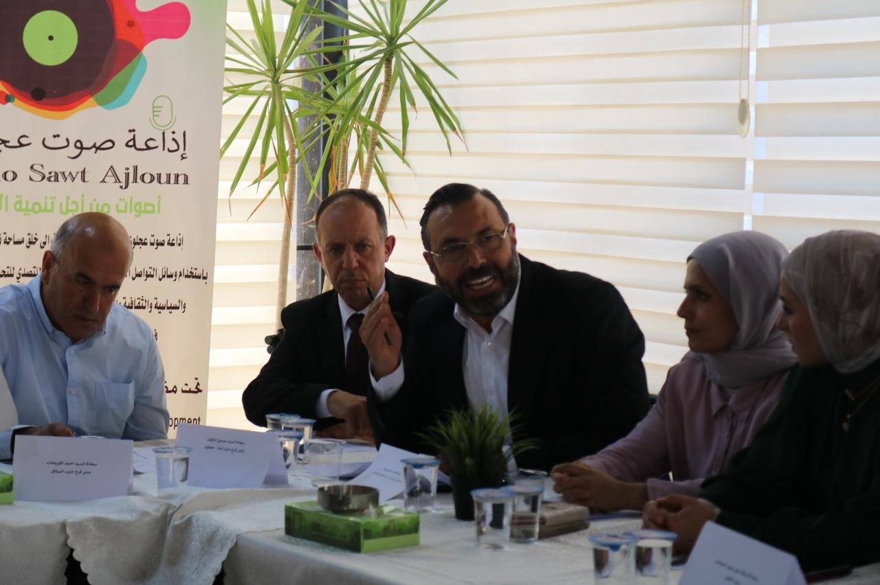 Panel of professionals seated at a conference table, discussing with a banner in Arabic behind them and plants nearby.