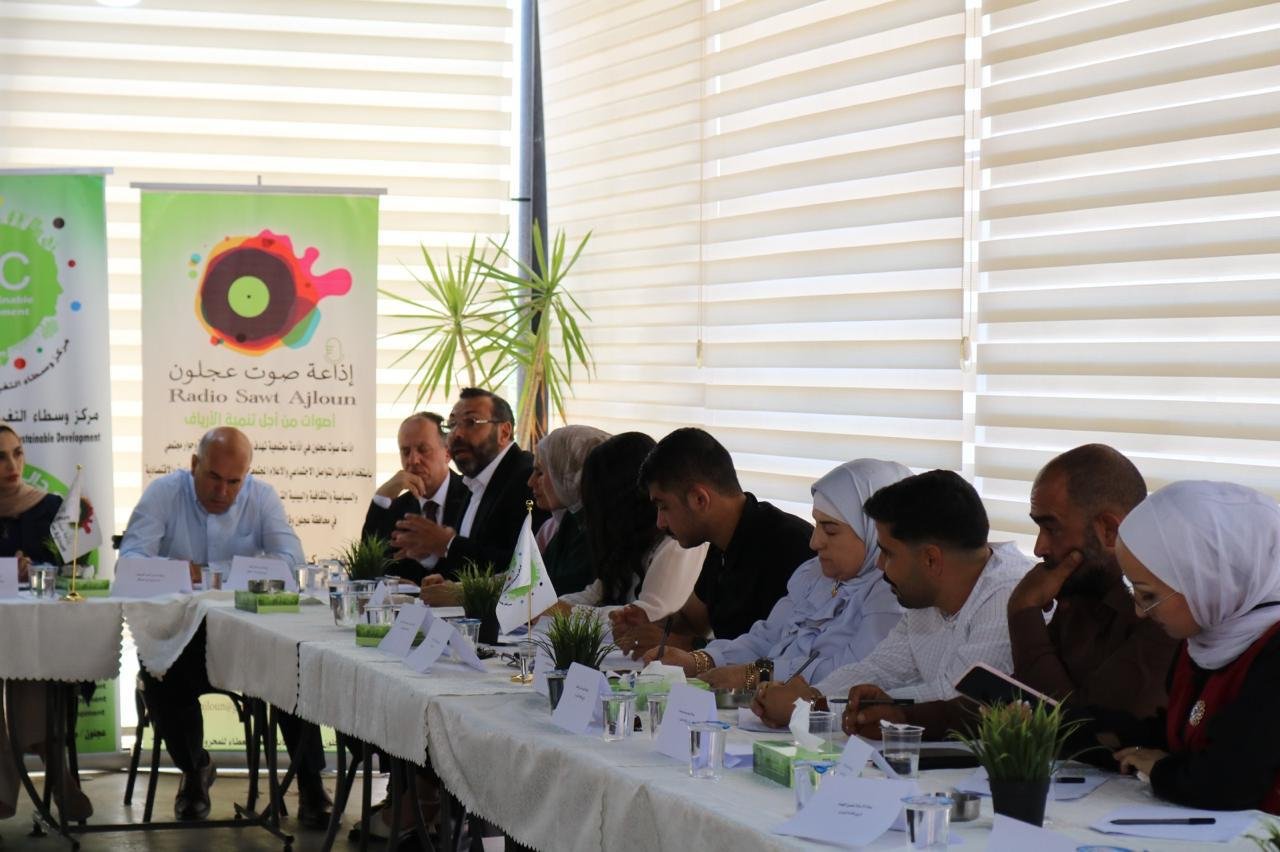 Group of diverse professionals seated around a long conference table, banners and plants in a bright room behind them.