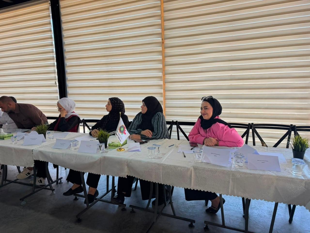 Panel of five people seated along a long table for a meeting, several women wearing hijabs, small plants and water glasses in front, and a flag at the center of the table.