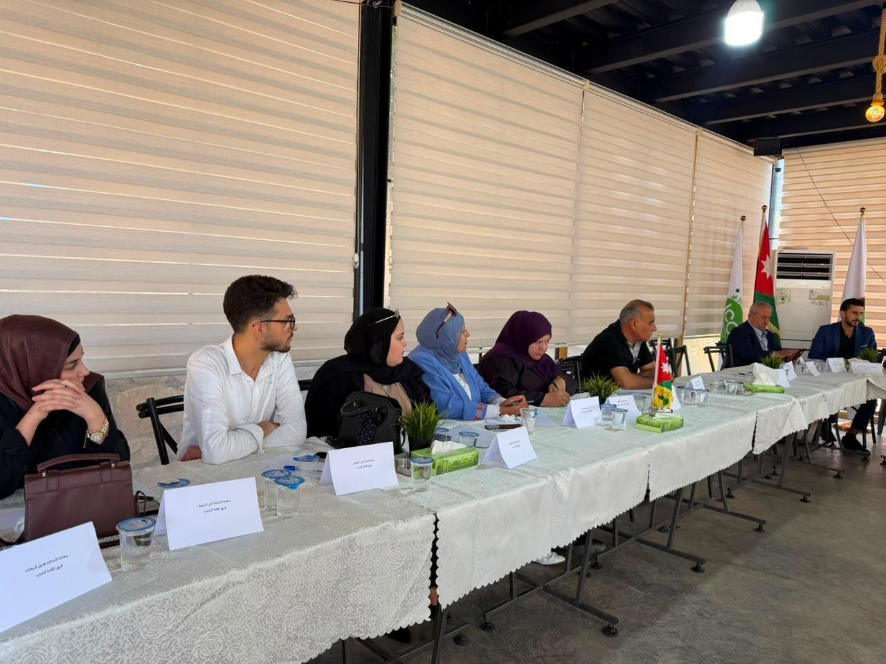 Diverse panel of men and women in hijabs seated along a long conference table with name cards and small plants at each place.